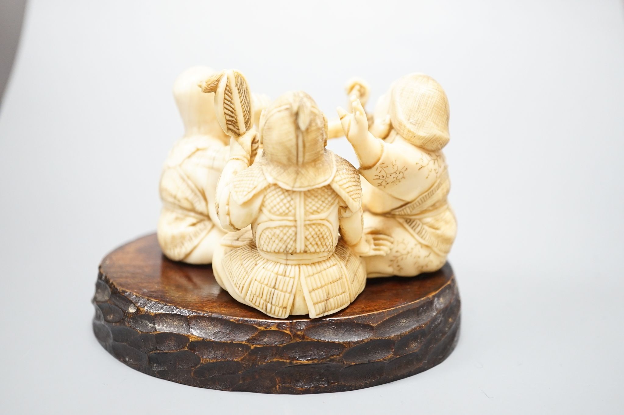 A Japanese ivory okimono of the gods Daikoku, Bishamonten and Fukurokuju, Meiji period, signed to a red lacquer tablet, 8cm wide, wood stand